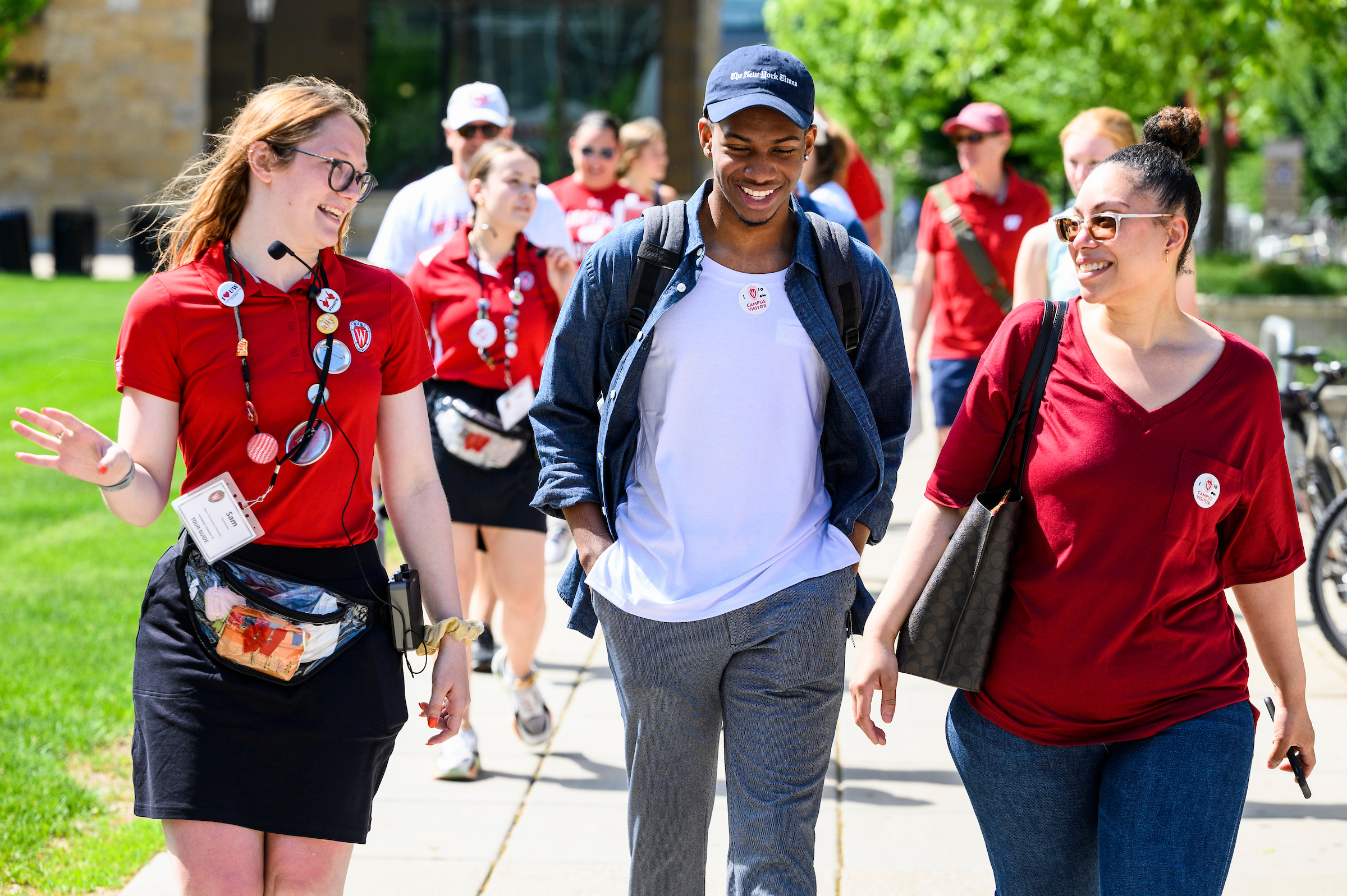 A male student in a blue baseball cap walks between two female staff members wearing red Wisconsin shirts.
